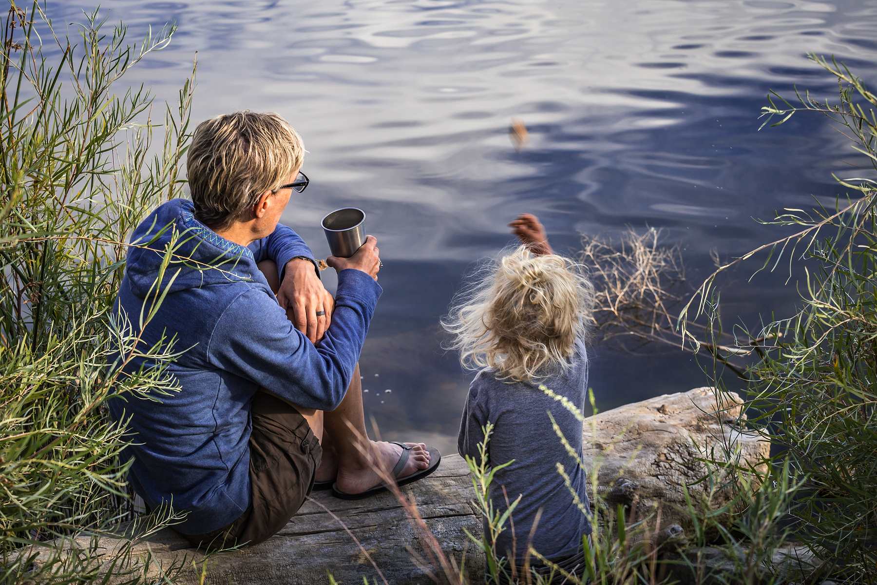 Older woman and a young child sitting on a log outside by the water.